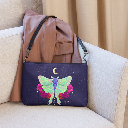 A navy blue clutch bag with a colorful moon moth and pink roses design, styled on a beige armchair with a brown jacket draped over it.