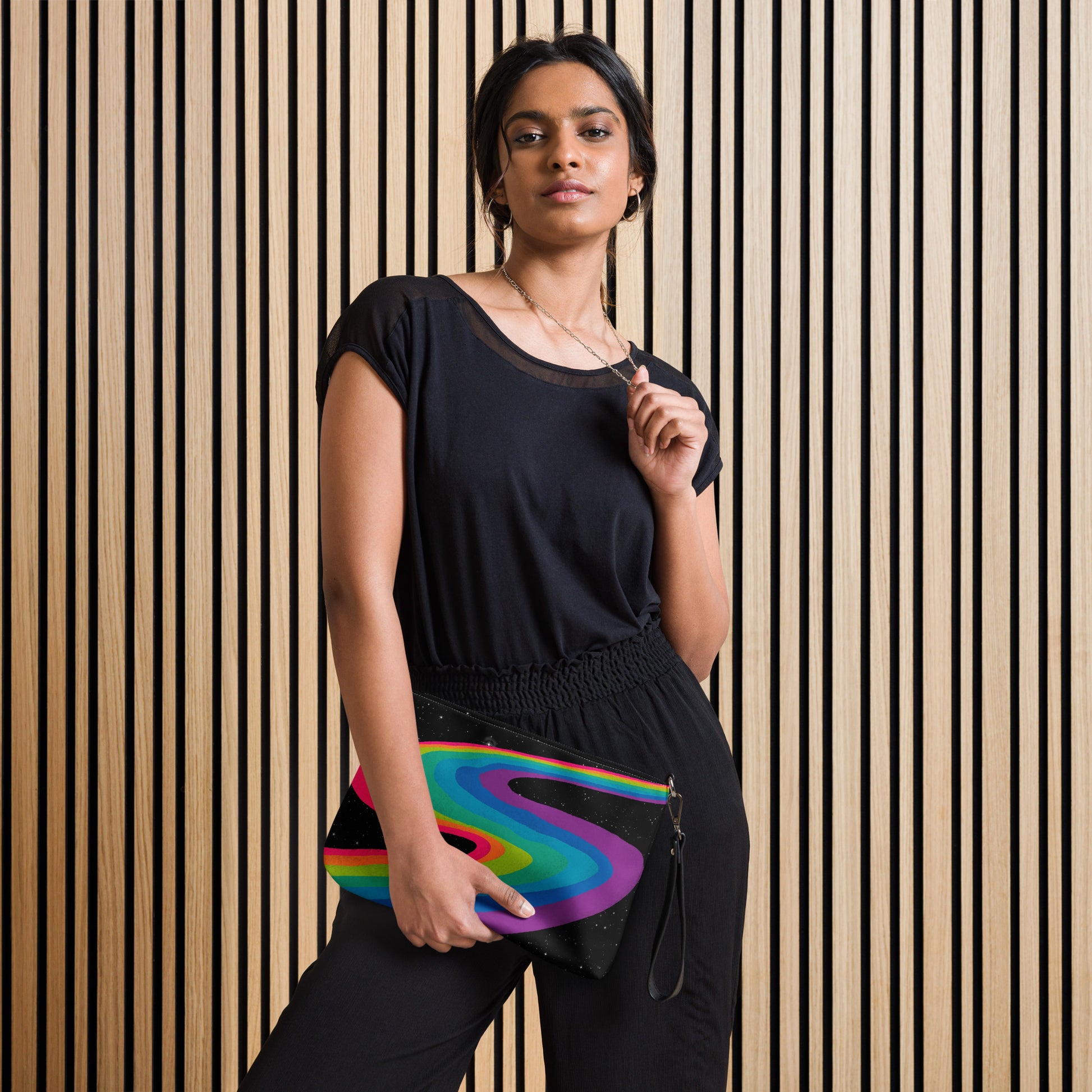 A model holding the Cosmic Rainbow Drift Crossbody Bag in black - hand-drawn swirling rainbow art on a starry galaxy background, faux leather with wrist strap, front view.