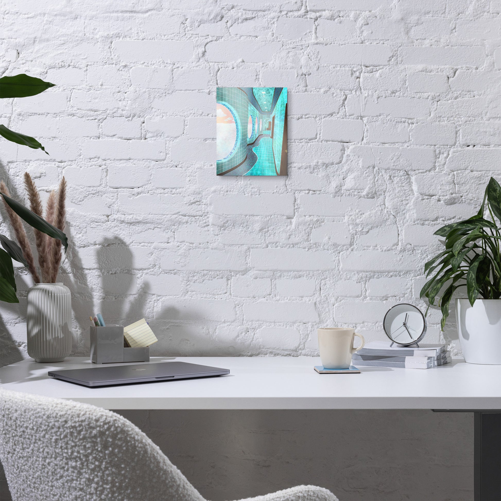 A small abstract wall art print with turquoise, teal, and cream tones hangs on a white brick wall above a styled home office desk with a laptop, mug, clock, and plants.